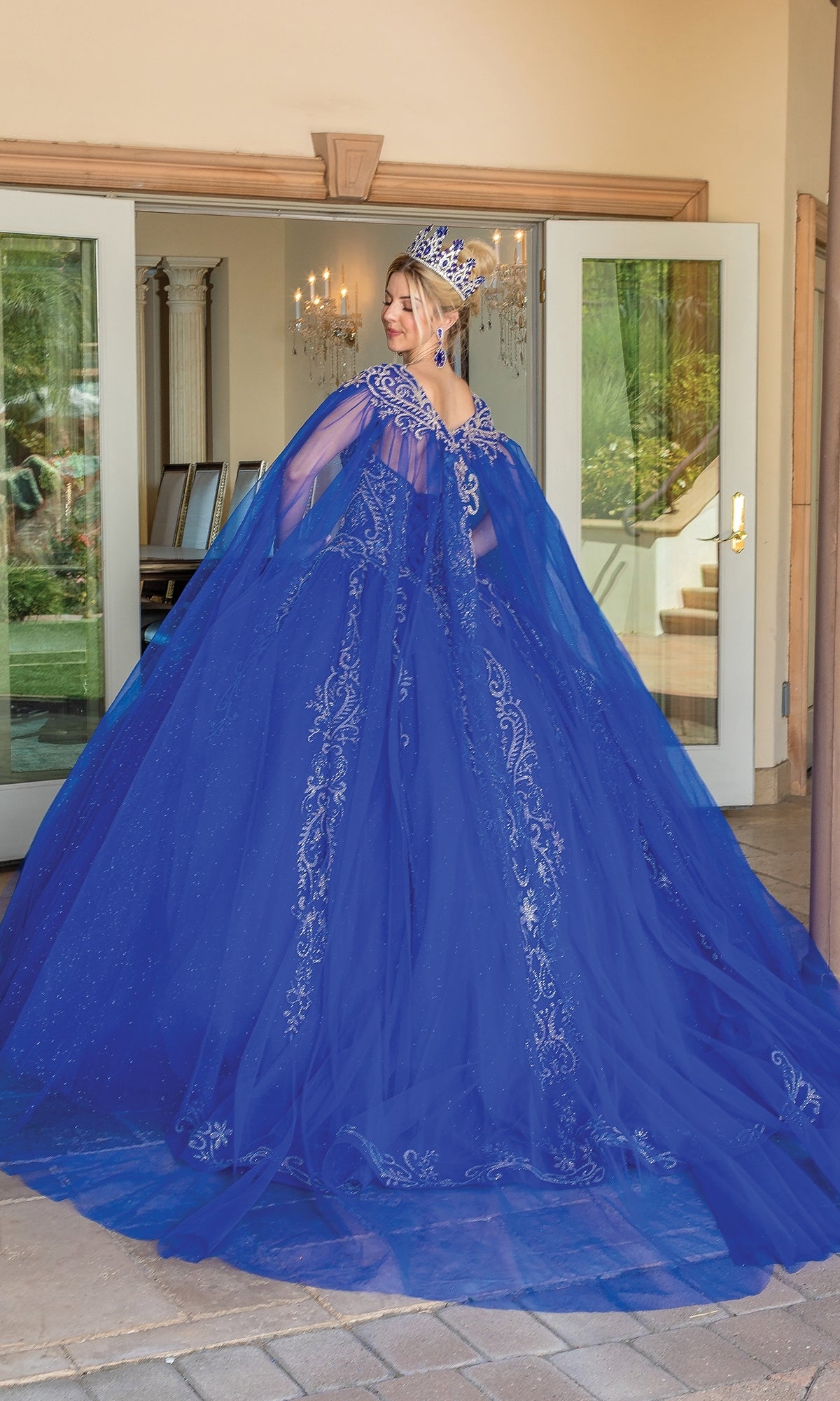 A woman wearing the Traditional Quinceañera Dress: Dancing Queen 1672 and a crown stands in front of open glass doors, looking over her shoulder.